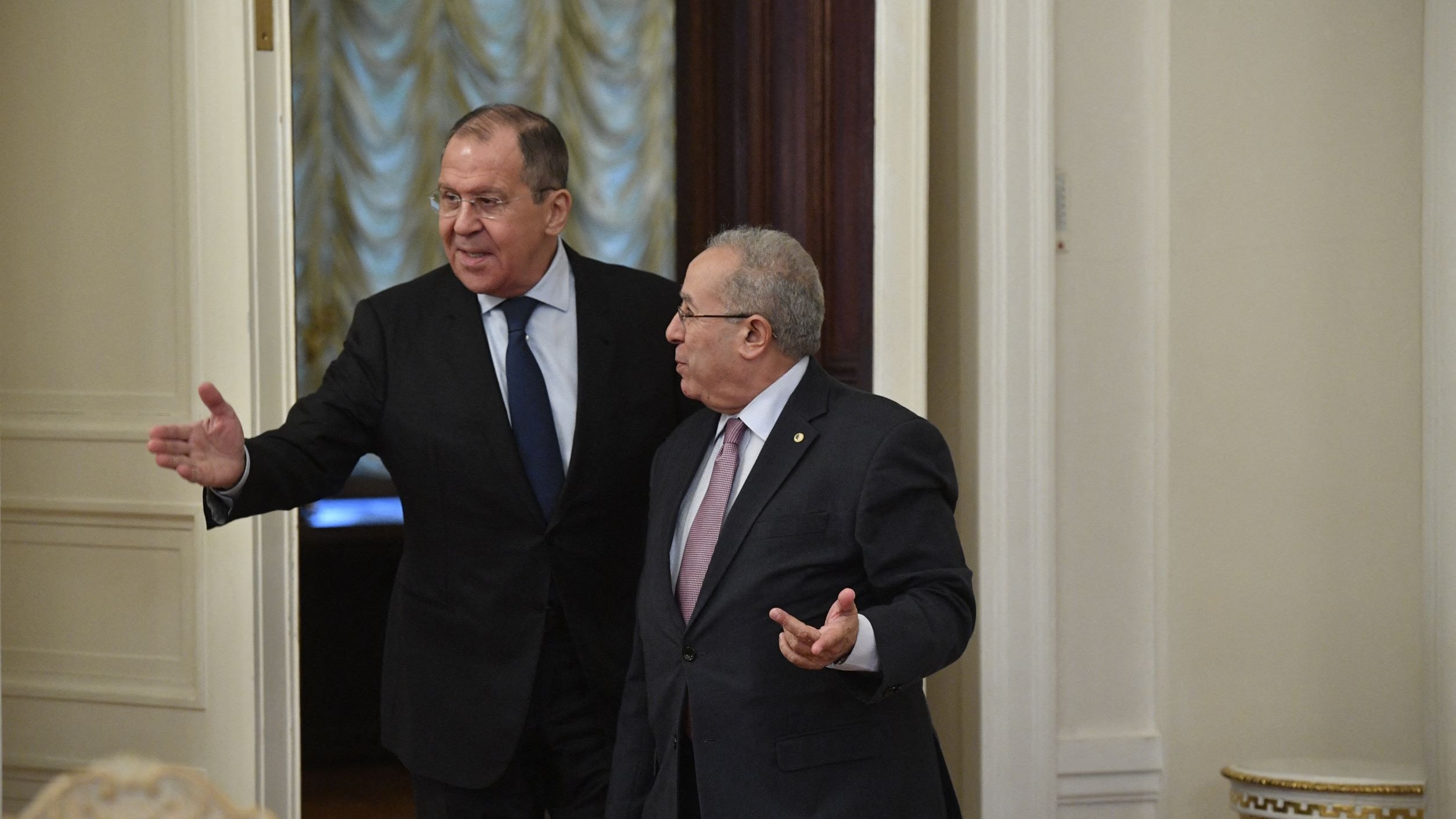 The image shows two men in a formal setting, likely inside a government building. One man is gesturing with his hand as he walks, indicating he is speaking or inviting the other man to follow. The second man appears to be listening attentively. Both are dressed in formal attire, suggesting a diplomatic or official meeting. The room has a sophisticated decor, with elements such as elegant walls and furniture visible in the background.
