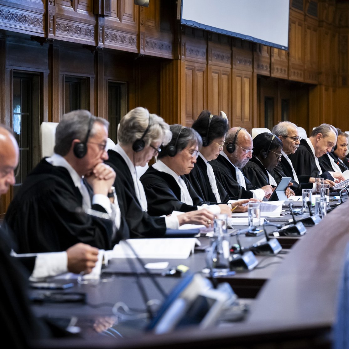 تصویر گروهی از قضات را نشان می‌دهد که در یک جلسه رسمی نشسته‌اند. آن‌ها لباس‌های مشکی قضائی به تن دارند و بسیاری از آن‌ها هدفون‌هایی بر سر دارند. سالن دارای دکوراسیون چوبی و شکلی کلاسیک است و یک میز بزرگ به شکل نیم‌دایره در مرکز تصویر وجود دارد. قضات در حال بحث یا مطالعه اسناد هستند و فضای جدی و رسمی حاکم بر جلسه احساس می‌شود.