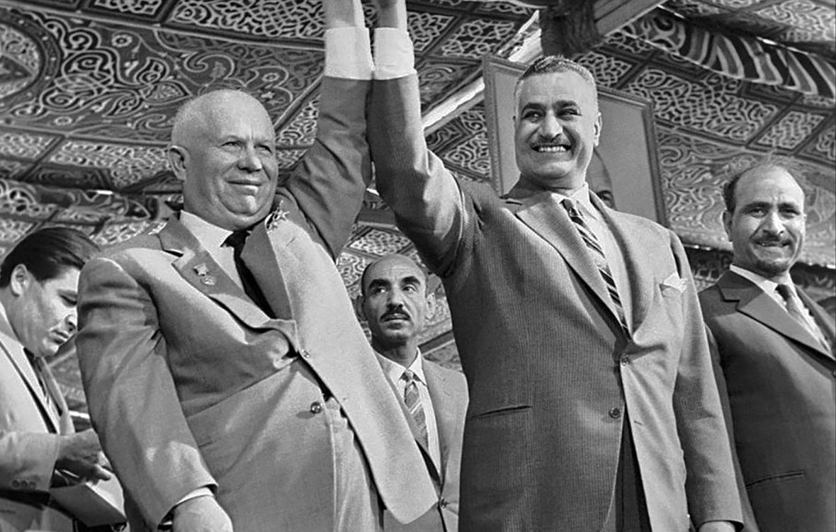 تظهر في الصورة مشهد تاريخي يجمع بين شخصين بارزين، حيث يحتفلان برفع أيديهما معًا تعبيرًا عن التضامن. الخلفية تبرز تفاصيل مميزة تشير إلى بيئة احتفالية، مع وجود عدد من الرجال الآخرين في الخلفية. يعكس هذا الموقف روح التعاون بين الشخصيتين في سياق تاريخي معين، مما يدل على أهمية اللحظة التي تم التقاطها فيها.