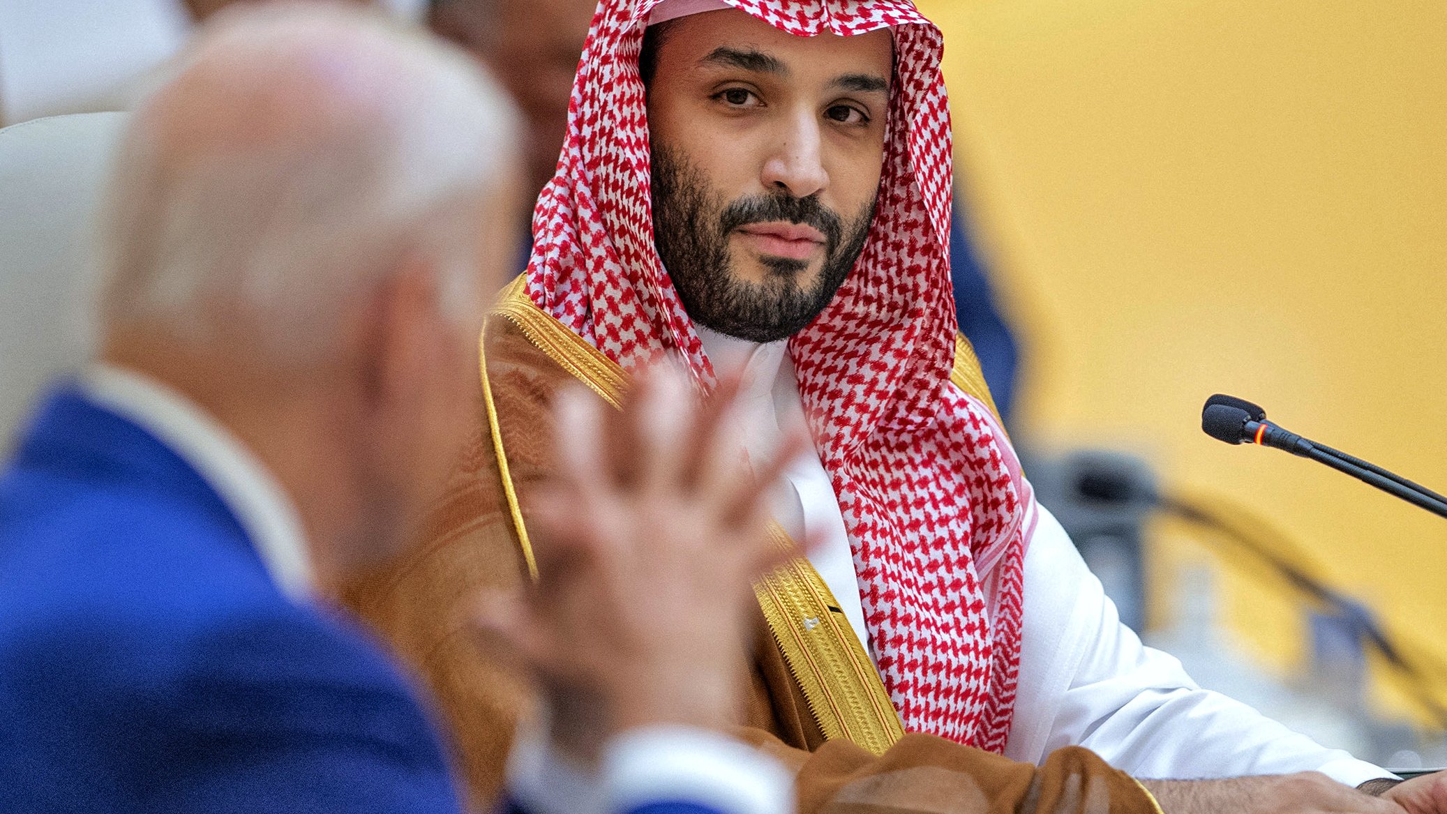 این تصویر مربوط به یک نشست یا جلسه رسمی است. در سمت چپ، یک مرد با لباس رسمی و کت و شلوار آبی نشسته است که به نظر می‌رسد در حال صحبت کردن است. در سمت راست، یک مرد دیگر با لباس سنتی عربی و عمامه قرمز و سفید، در حال گوش دادن به سخنان فرد مقابل است. پس‌زمینه سالن جلسه به طرزی طراحی شده که نشان‌دهنده یک محیط رسمی و دیپلماتیک است.