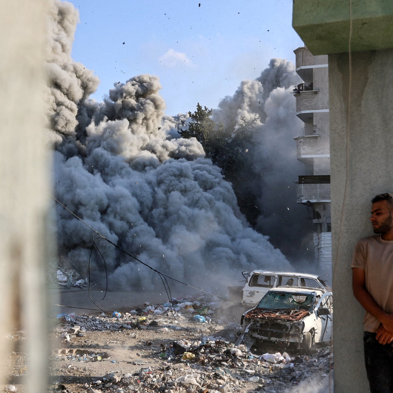 Fumi grigi salgono da un'esplosione, un uomo appoggiato a un pilastro vicino a un'auto distrutta.