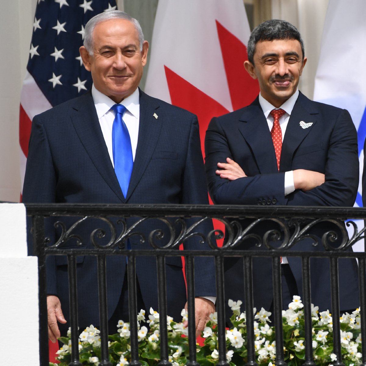 L'image montre trois hommes debout sur un balcon, avec des drapeaux en arrière-plan, dont ceux des États-Unis, du Canada, et d'Israël. Les hommes semblent souriants et détendus, portant des costumes formels. Il y a des fleurs blanches qui décorent le balcon. La scène exprime une ambiance de diplomatie ou de réunion internationale.