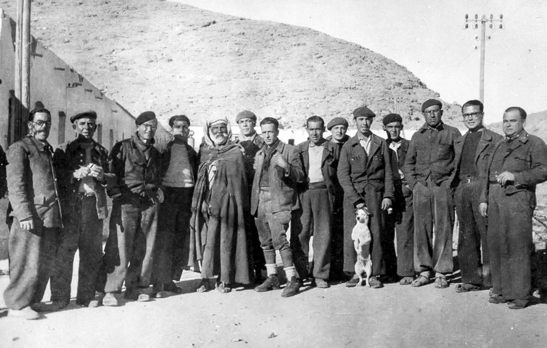 La imagen muestra un grupo de hombres de pie en un entorno rural, posiblemente en una zona montañosa. Están vestidos con ropas tradicionales y de trabajo, algunos con boinas. Hay también un perro que se encuentra entre ellos. La atmósfera es de camaradería, y el paisaje detrás sugiere una época pasada, posiblemente de principios del siglo XX. Los rostros reflejan diferentes expresiones, mientras que la composición transmite una sensación de unión entre los miembros del grupo.