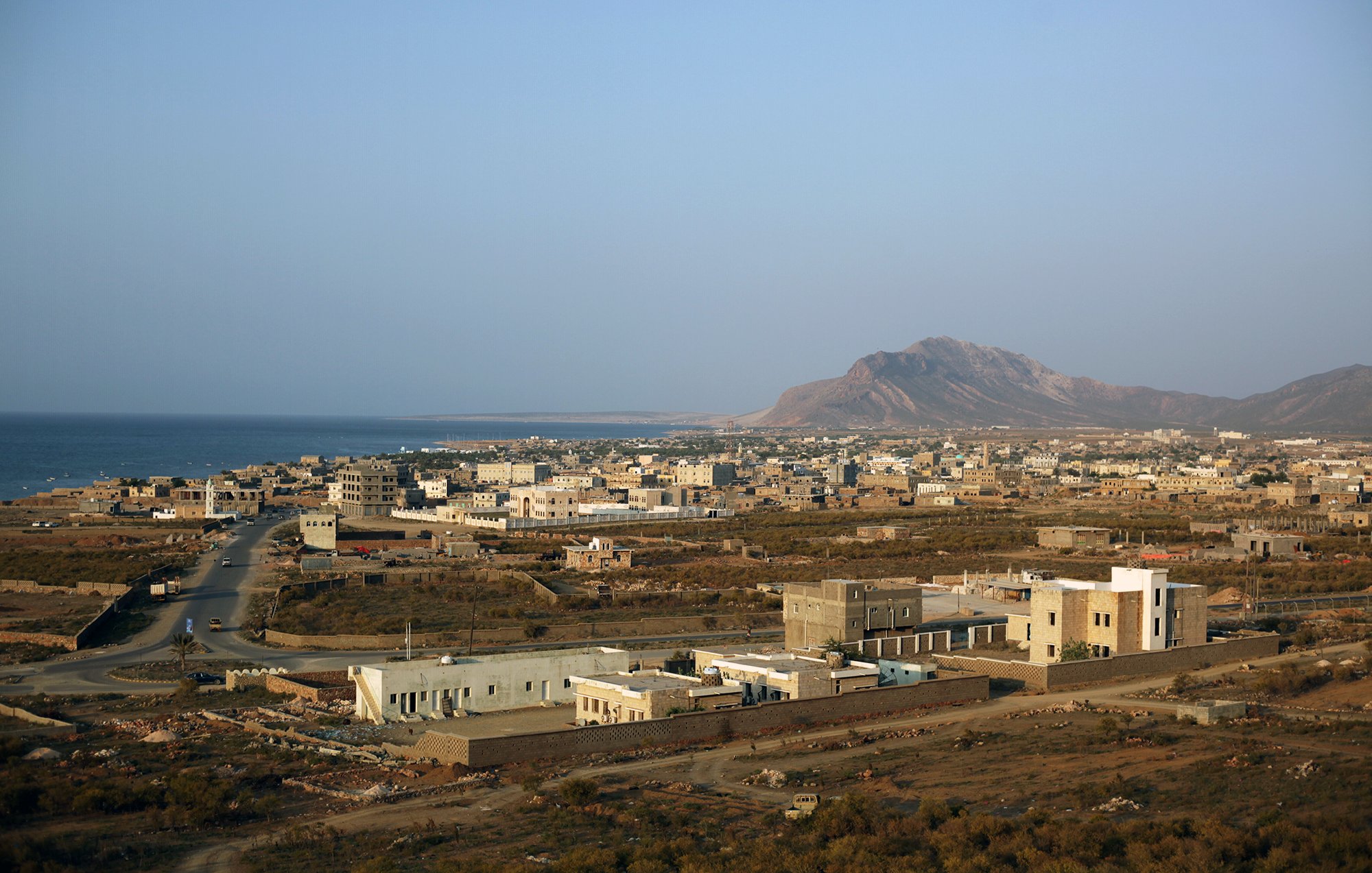 تظهر الصورة منظرًا خلابًا لبلدة ساحلية تطل على البحر. يُظهر المشهد منازل مبنية بشكل متفرق، بعضها حديث والآخر تقليدي، مع شوارع تمتد بين المباني. في الخلفية، يمكن رؤية سلسلة جبال، بينما يمتد البحر الفسيح على الجهة اليسرى. السماء صافية وتبدو أشعة الشمس مشرقة، مما يضفي شعورًا بالهدوء والجمال الطبيعي على المكان.