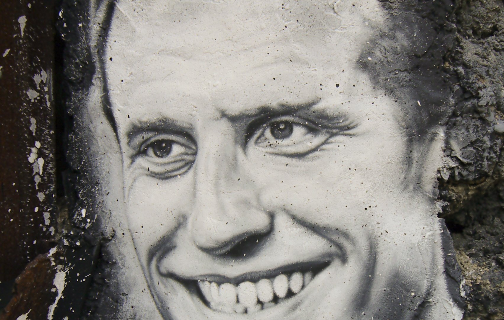 The image features a black-and-white mural of a smiling man. The artwork is set against a textured, stone wall, which adds depth to the scene. The man's expression conveys warmth and friendliness, with a prominent smile and bright eyes. The details are finely rendered, showcasing the artist's skill in capturing facial features and expressions.