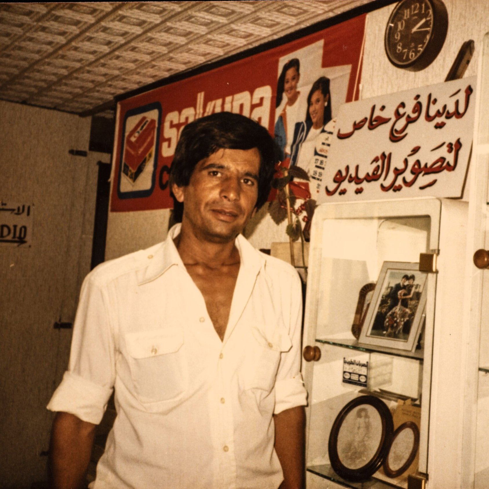 L'immagine mostra un uomo in un ambiente che sembra essere un negozio o un'esposizione. Indossa una camicia bianca e ha i capelli scuri e lunghi. Sullo sfondo, si possono vedere vari oggetti esposti, tra cui quadri e cornici. C'è anche un grande cartello con del testo in arabo e un orologio visibile. L'atmosfera complessiva trasmette un senso di nostalgia.