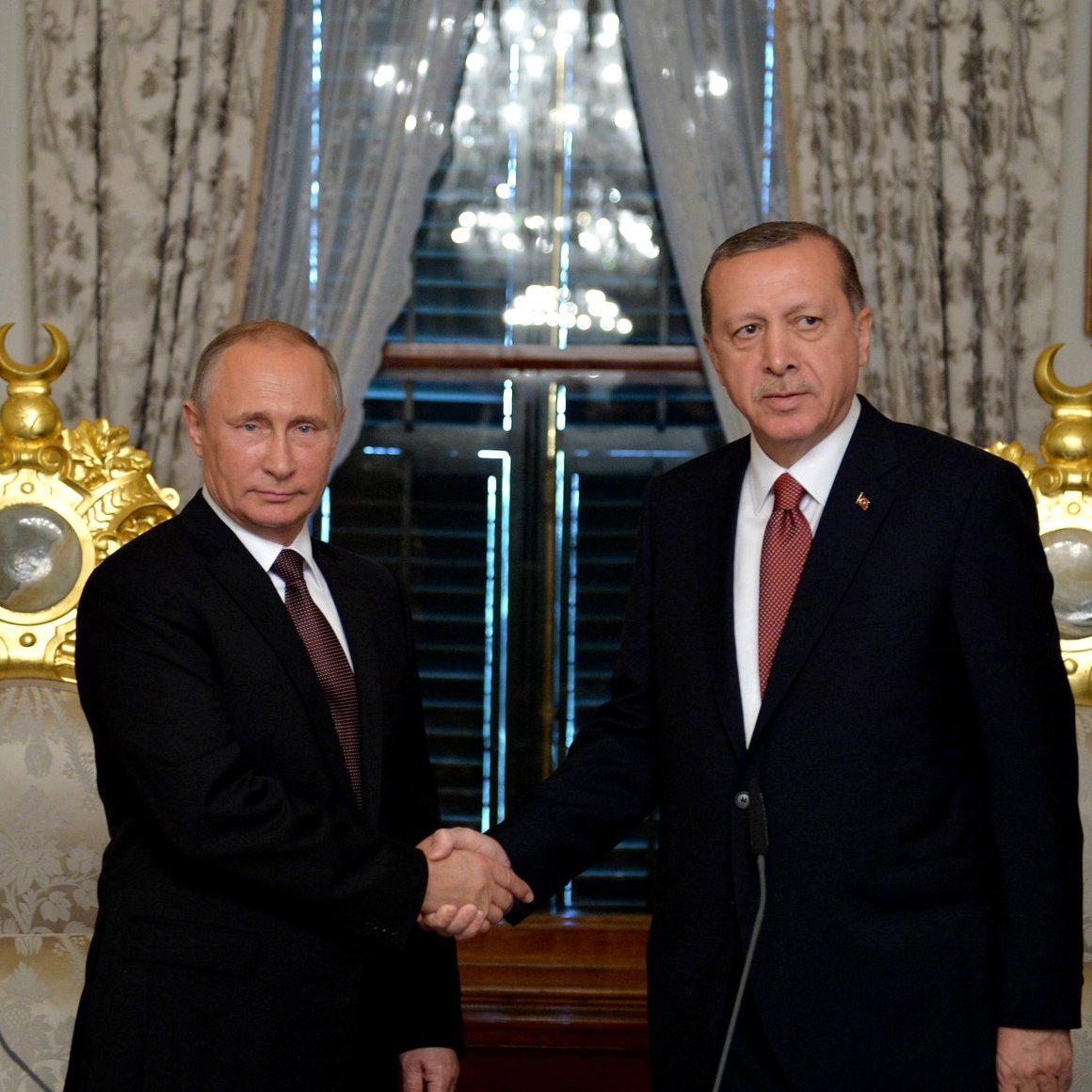 En la imagen se pueden ver a dos líderes políticos, uno de Rusia y otro de Turquía, dándose la mano en un acto formal. Ambos están vestidos con trajes oscuros y corbatas, y parecen estar en un entorno elegante, rodeados de decoraciones doradas y banderas de sus respectivos países. La atmósfera sugiere un momento de colaboración o acuerdo entre las dos naciones.