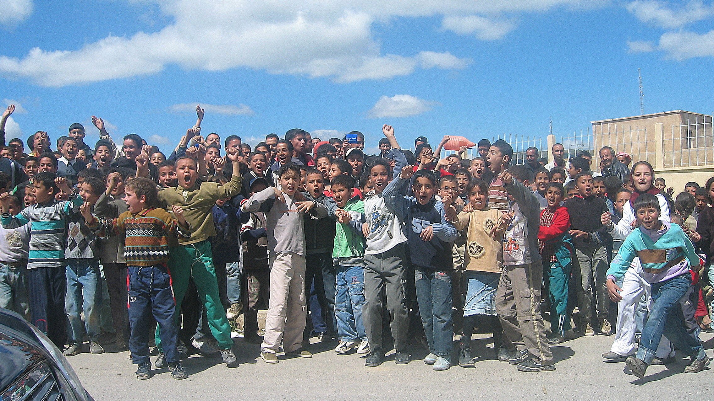L'image montre un groupe important d'enfants et d'adolescents rassemblés en plein air. Ils semblent joyeux et excités, levant les bras et faisant des gestes. Le ciel est clair avec quelques nuages, et l'environnement urbain en arrière-plan suggère un cadre scolaire ou communautaire. Les enfants portent des vêtements variés, reflet de leur diversité. L'atmosphère semble animée et festive.