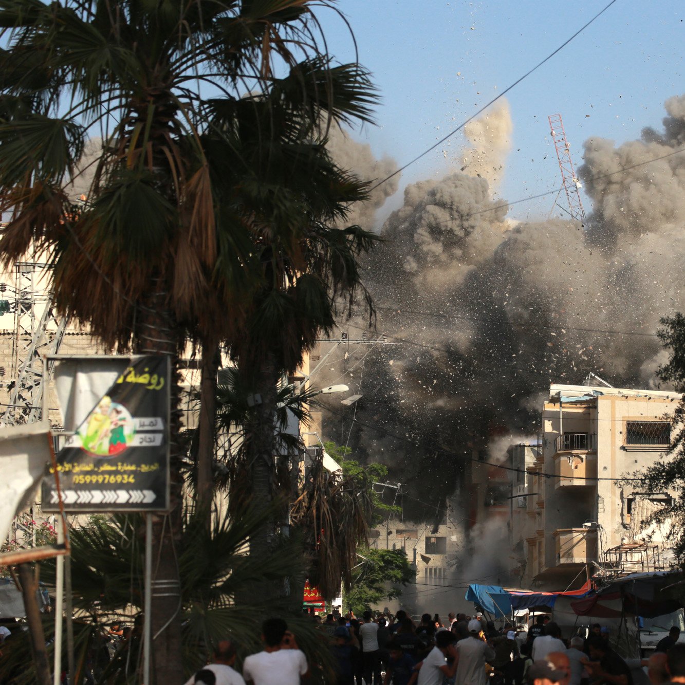 La imagen muestra una escena de destrucción y caos en una ciudad. Se puede ver una gran nube de polvo y escombros elevándose en el aire, lo que sugiere que ha ocurrido una explosión o un colapso de un edificio. En primer plano, hay un grupo de personas que parecen estar observando la situación, algunas de ellas con expresiones de sorpresa o preocupación. Alrededor, hay palmeras y estructuras urbanas que indican que se trata de un área urbana. El cielo es de un color azul claro, contrastando con el humo gris.