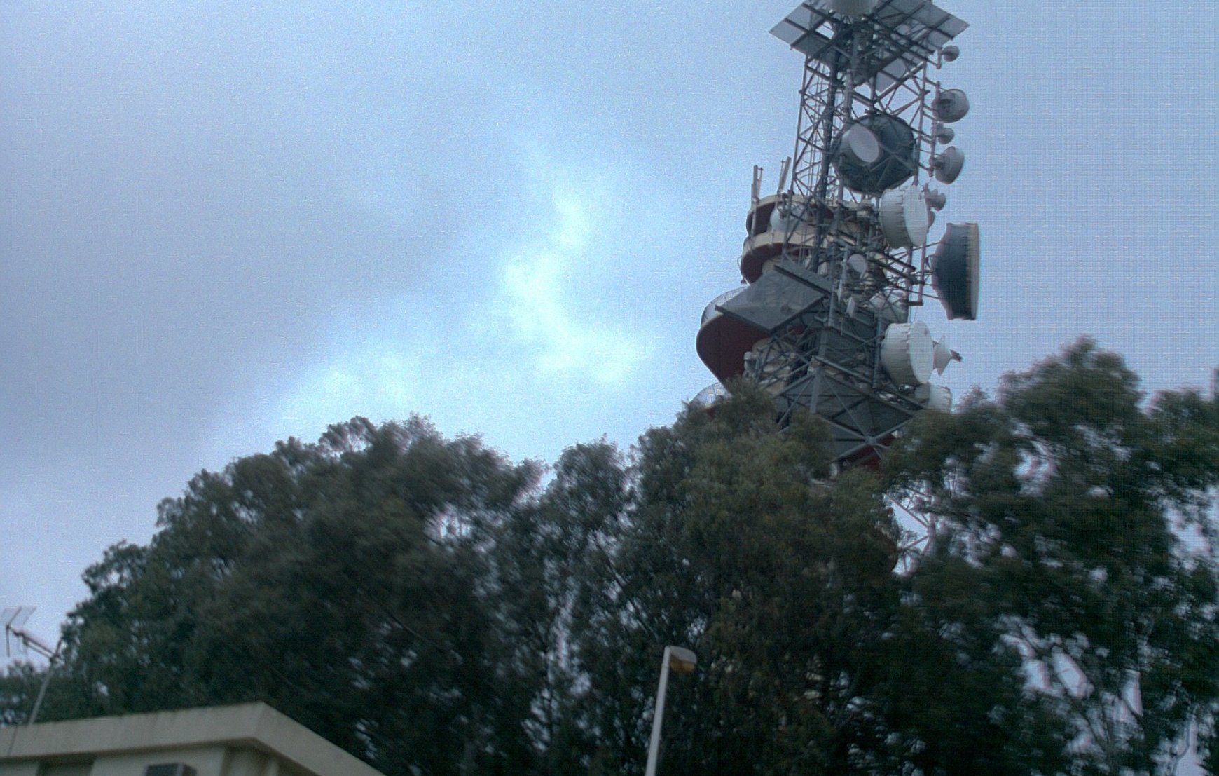 L'image montre une tour de télécommunication élevée, dotée de plusieurs antennes et équipements en haut. Elle est entourée de grands arbres verts et se dresse à côté d'un bâtiment plus petit, probablement un centre d'opérations ou de maintenance. Le ciel est nuageux, ce qui donne une ambiance légèrement grise et sombre à la scène.