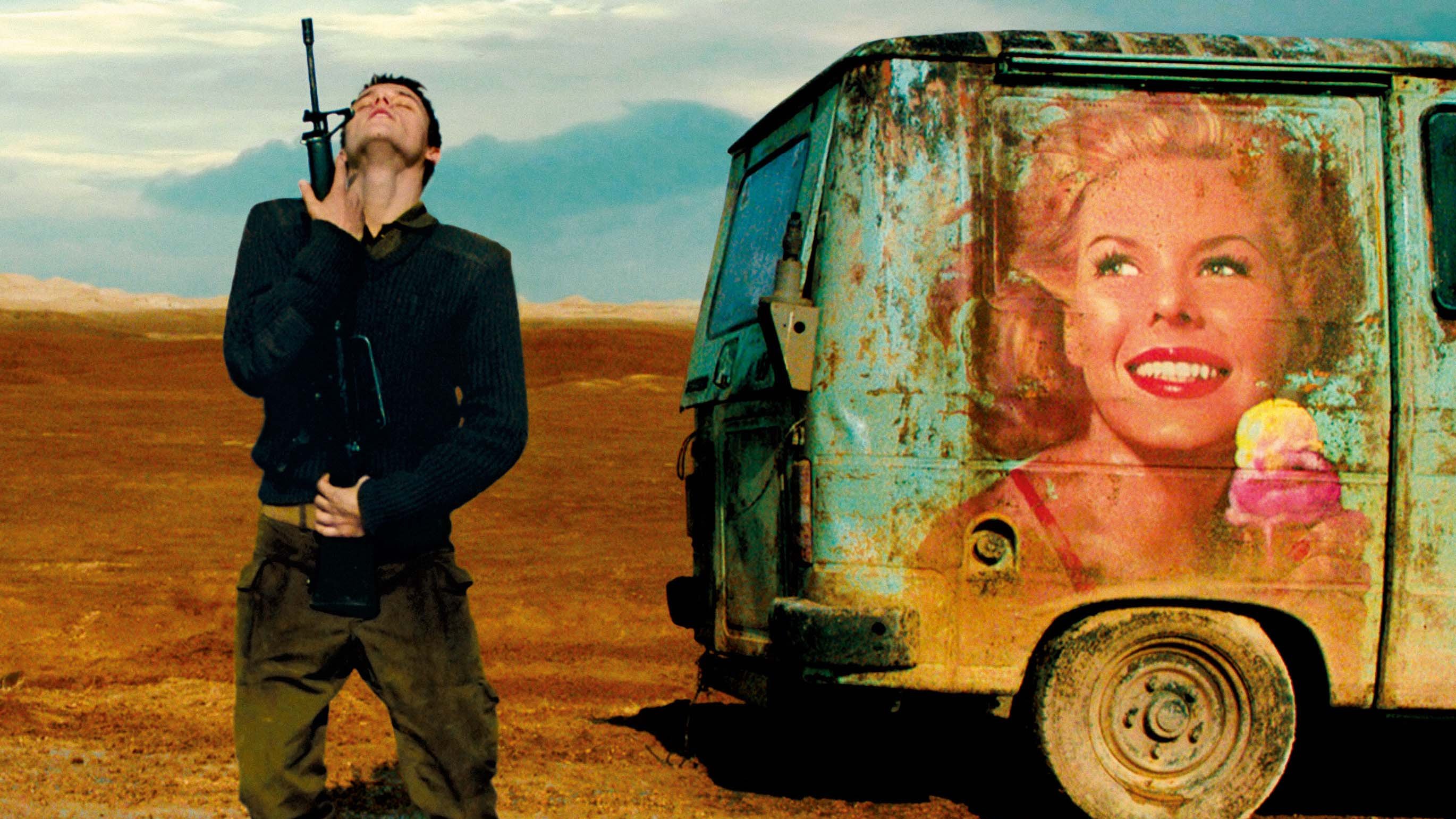 L'image montre un homme debout dans un paysage désertique, avec une expression qui semble mélancolique ou contemplative. Il porte une arme à feu à la main et est habillé de manière militaire. À côté de lui se trouve un vieux camion, sur lequel est peint un portrait coloré d'une femme souriante tenant une glace. Le contraste entre l'aspect rustique du véhicule et l'image joyeuse de la femme crée une atmosphère intrigante, mêlant des éléments de décor et d'émotion. Le ciel, quant à lui, est nuageux, ajoutant une dimension dramatique à la scène.