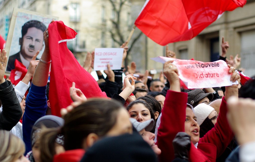 L'image montre une foule de manifestants rassemblés, brandissant des drapeaux rouges et des pancartes. Certains visages sont masqués, tandis que d'autres expriment des émotions intenses, comme la détermination ou la colère. On aperçoit aussi un portrait d'une figure emblématique en arrière-plan. Les slogans sur les pancartes semblent appeler au changement et à la liberté, indiquant un contexte de protestation politique. L'atmosphère est chargée d'énergie collective.