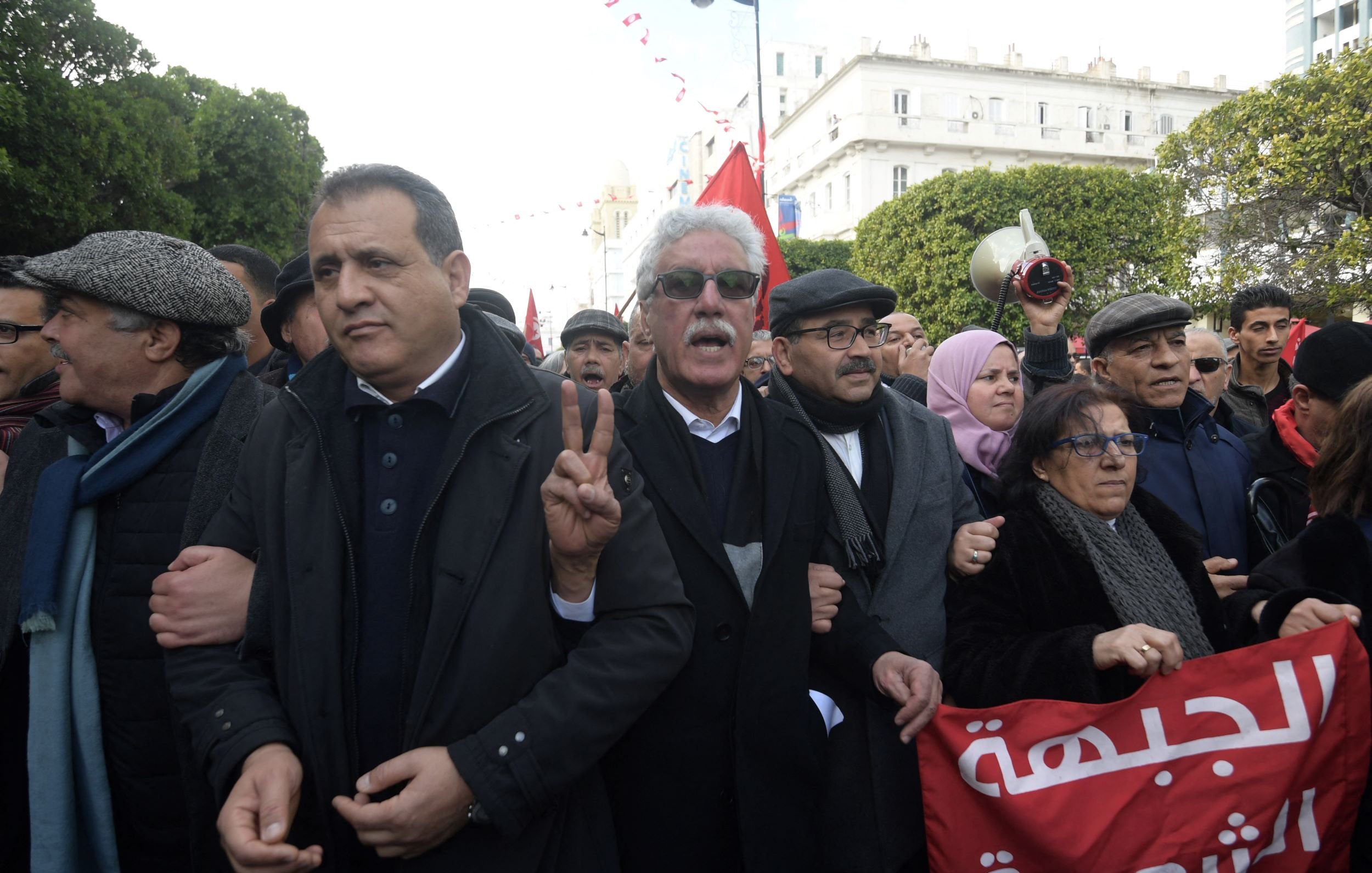 L'immagine mostra un gruppo di persone in una manifestazione. Alcuni uomini e donne stanno insieme, alcuni con espressioni decise e altri sorridenti. Alcuni partecipanti indossano cappelli e giacche scure. Una donna tiene un cartello redatto in arabo, mentre un altro uomo alza la mano formando il segno della vittoria. Sullo sfondo si vedono bandiere e altri manifestanti, creando un'atmosfera di mobilitazione e unità.
