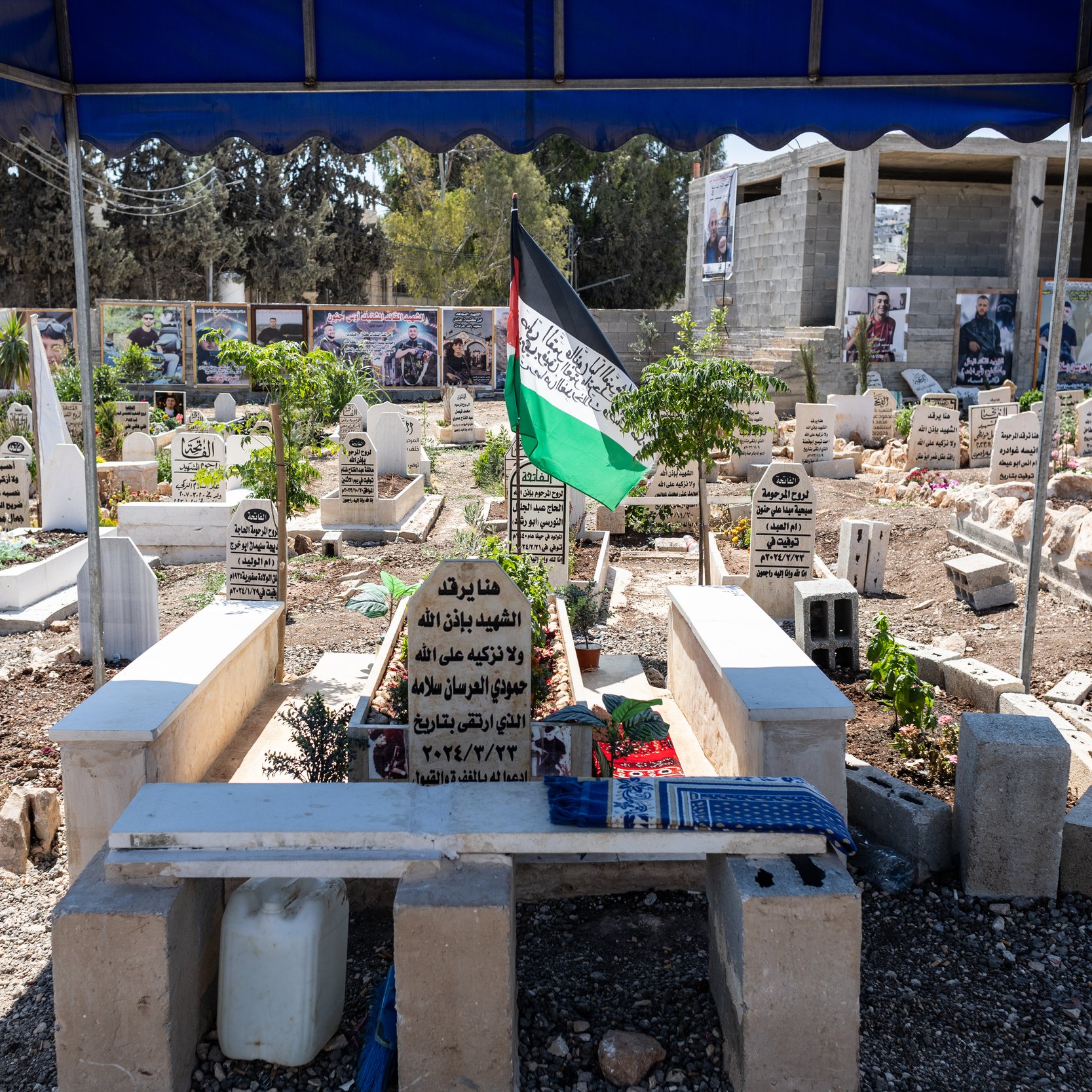 L'immagine mostra un cimitero all'aperto, con diverse tombe disposte in un'area ben curata. Le lapidi sono in varie posizioni e materiali, molte sono contrassegnate con iscrizioni in arabo. Ci sono anche alcune piante verdi e fiori, che aggiungono un tocco di vita al luogo. Al centro, si può vedere una bandiera con i colori della Palestina che sventola, e sullo sfondo ci sono delle fotografie appese, probabilmente in memoria di persone care. Il cimitero è coperto da una tettoia, creando un'area protetta. L'atmosfera è solenne e rispettosa.