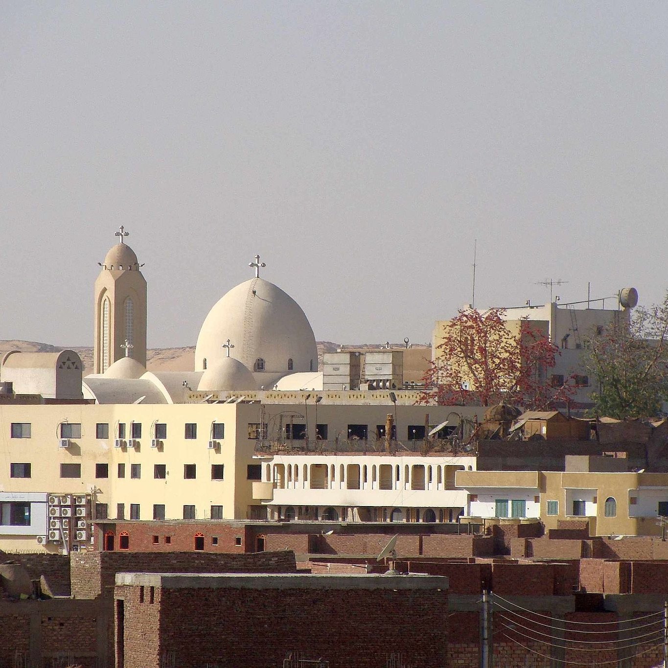 الصورة تظهر مجموعة من المباني في منطقة حضرية، حيث يتواجد مباني ذات طراز معماري مميز، بما في ذلك الكنائس التي تتضمن قبابًا وجرسًا. الخلفية تحتوي على تلال وصحراء، مما يوحي بأن المنطقة قد تكون قريبة من الطبيعة. يوجد أيضًا مجموعة من المباني السكنية المنخفضة، تظهر على اليمين، مما يعطي انطباعًا عن الحياة اليومية في تلك المنطقة.
