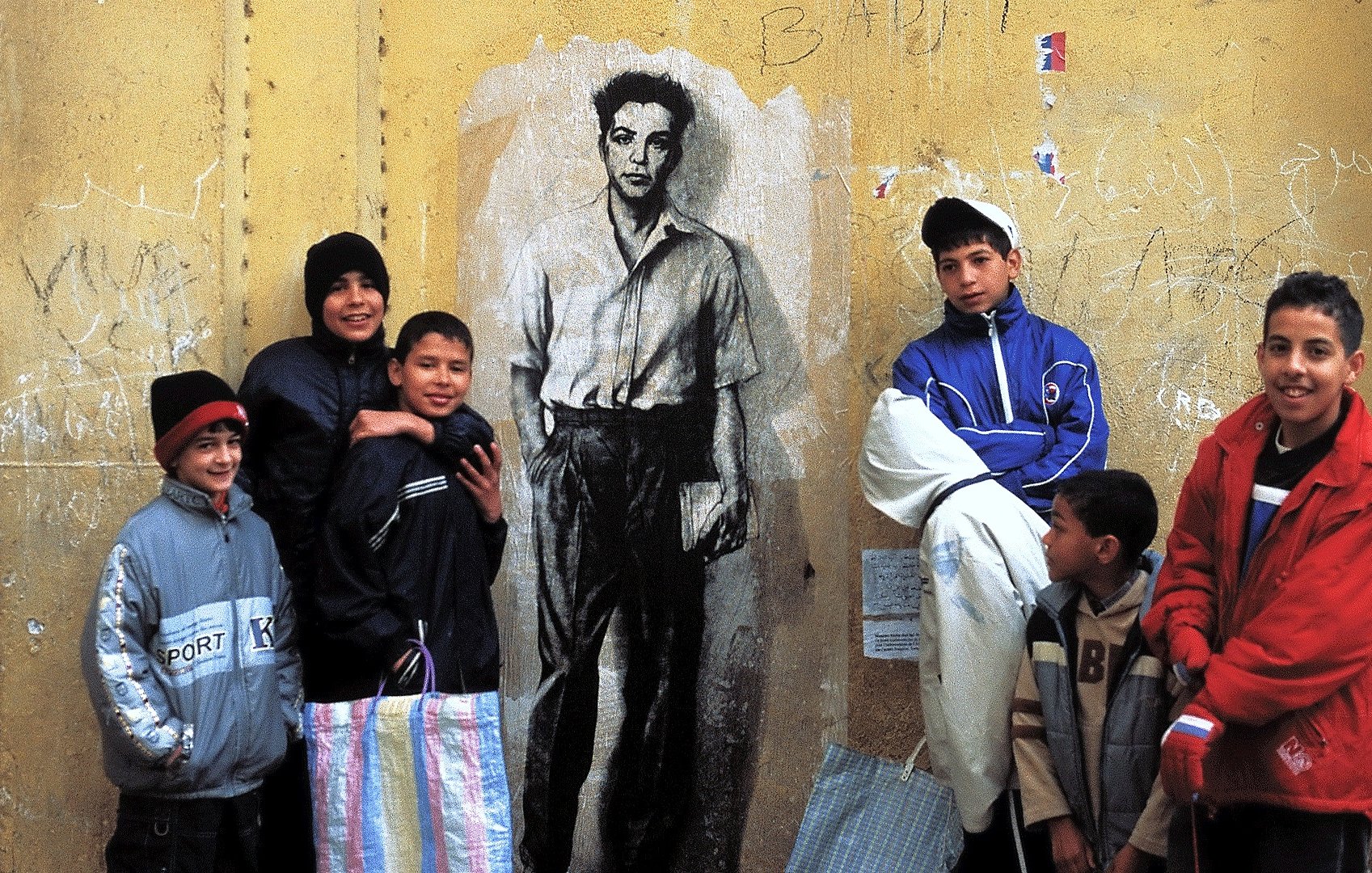 La imagen muestra a un grupo de niños posando al frente de una pared amarilla en la que hay un mural de un hombre en blanco y negro. Los niños parecen estar disfrutando del momento y están vestidos con ropa de abrigo. Algunos de ellos sostienen bolsas, posiblemente de tela. La escena transmite una sensación de comunidad y alegría, en contraste con el fondo urbano y desgastado de la pared.