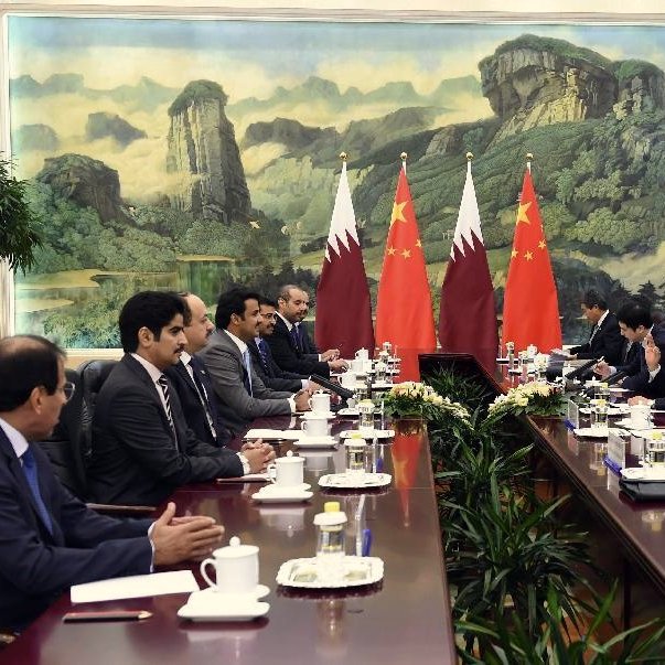 L'image montre une réunion formelle dans une salle de conférence. On peut y voir plusieurs personnes assises autour d'une longue table. À l'arrière-plan, il y a deux drapeaux, celui du Qatar et celui de la Chine, ainsi qu'une grande fresque représentant des montagnes et des paysages. Les participants semblent engagés dans une discussion, avec des tasses et des documents disposés sur la table. L'ambiance semble sérieuse et professionnelle, indiquant un dialogue diplomatique entre les deux pays.