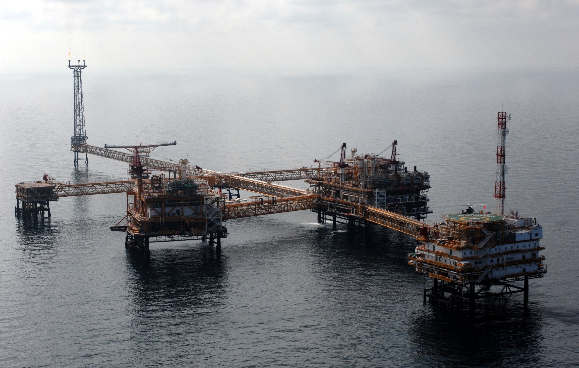 The image depicts an offshore oil platform complex situated in the ocean. The facility is characterized by several interconnected structures, platforms, and towers. These platforms likely house drilling equipment and machinery used for extracting oil or gas from beneath the seabed. The background features calm waters and a cloudy sky, suggesting a remote marine environment. The arrangement of the platforms indicates a significant operation for energy production.