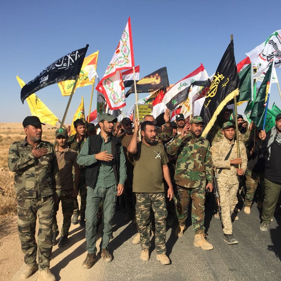 The image depicts a group of men participating in a march or rally, holding various flags. The men are dressed in military-style clothing and appear to be organized, with some wearing uniforms and others in casual attire. The flags they carry include a mix of colors and designs, suggesting they represent different factions or groups. The scene is set in an open area, likely with dry or arid terrain in the background, and the sky is clear. The atmosphere seems to indicate a demonstration or a show of solidarity among the participants.