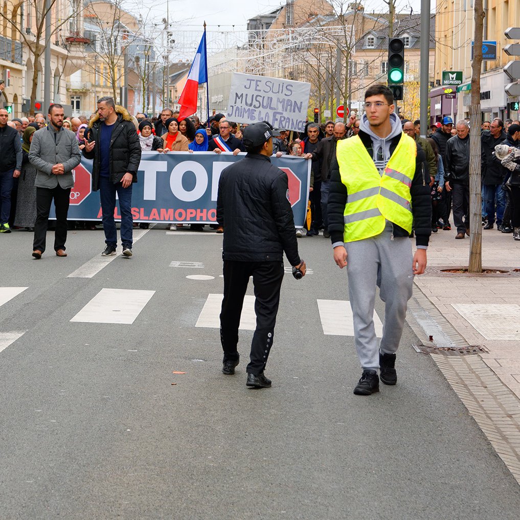 الصورة تظهر مجموعة من الناس يتظاهرون في شارع. في المقدمة، هناك شخصان يرتديان سترة عاكسة، ويبدو أنهما يجريان أو يقومان بمراقبة المظاهرة. في الخلفية، يمكن رؤية لافتة كبيرة مكتوب عليها "STOP ISLAMOPHOBIE"، مما يشير إلى أن المظاهرة تتعلق بمناهضة الإسلاموفوبيا. المتظاهرون يحملون لافتات ويبدو أنهم يتجمعون بشكل منظم. الجو العام يعكس النشاط والحيوية.