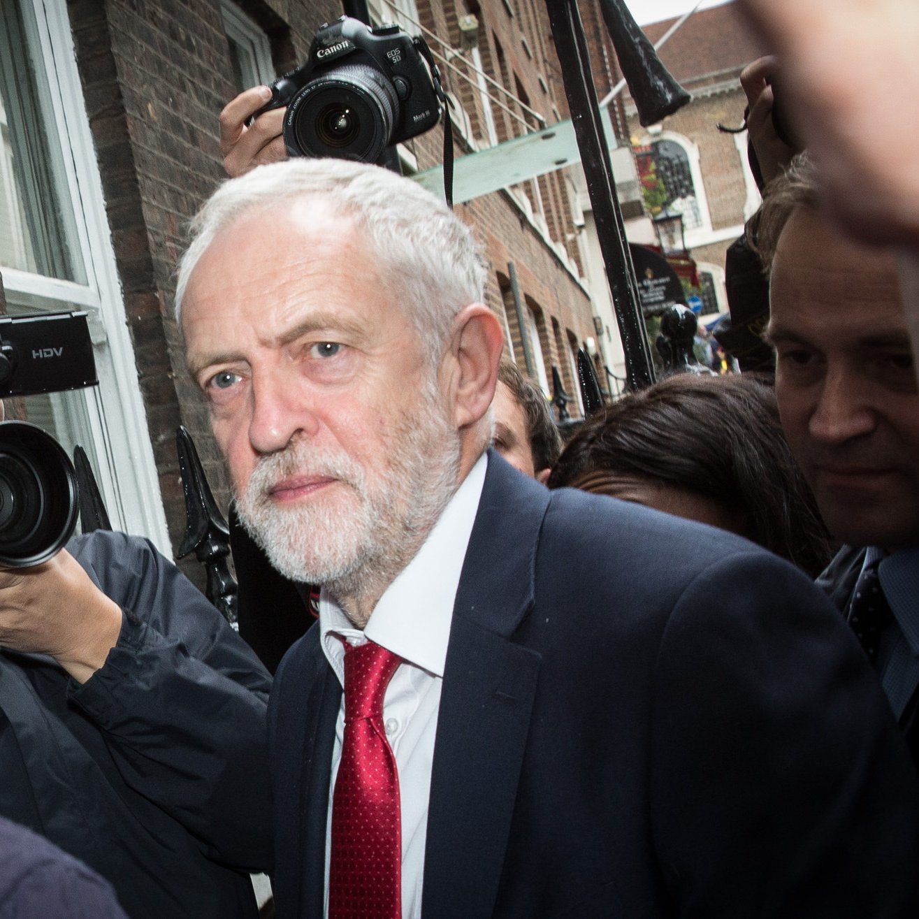 الصورة تظهر رجلًا متوسط العمر ذو لحية وشعر رمادي، يرتدي بدلة داكنة وربطة عنق حمراء. يبدو أنه في موقف يتطلب الانتباه، حيث تحيط به مجموعة من الكاميرات والصحفيين الذين يلتقطون صورًا له. الخلفية تعكس بيئة حضرية مع مبانٍ طوبية، وتعكس الحركة والاهتمام بالشخص.