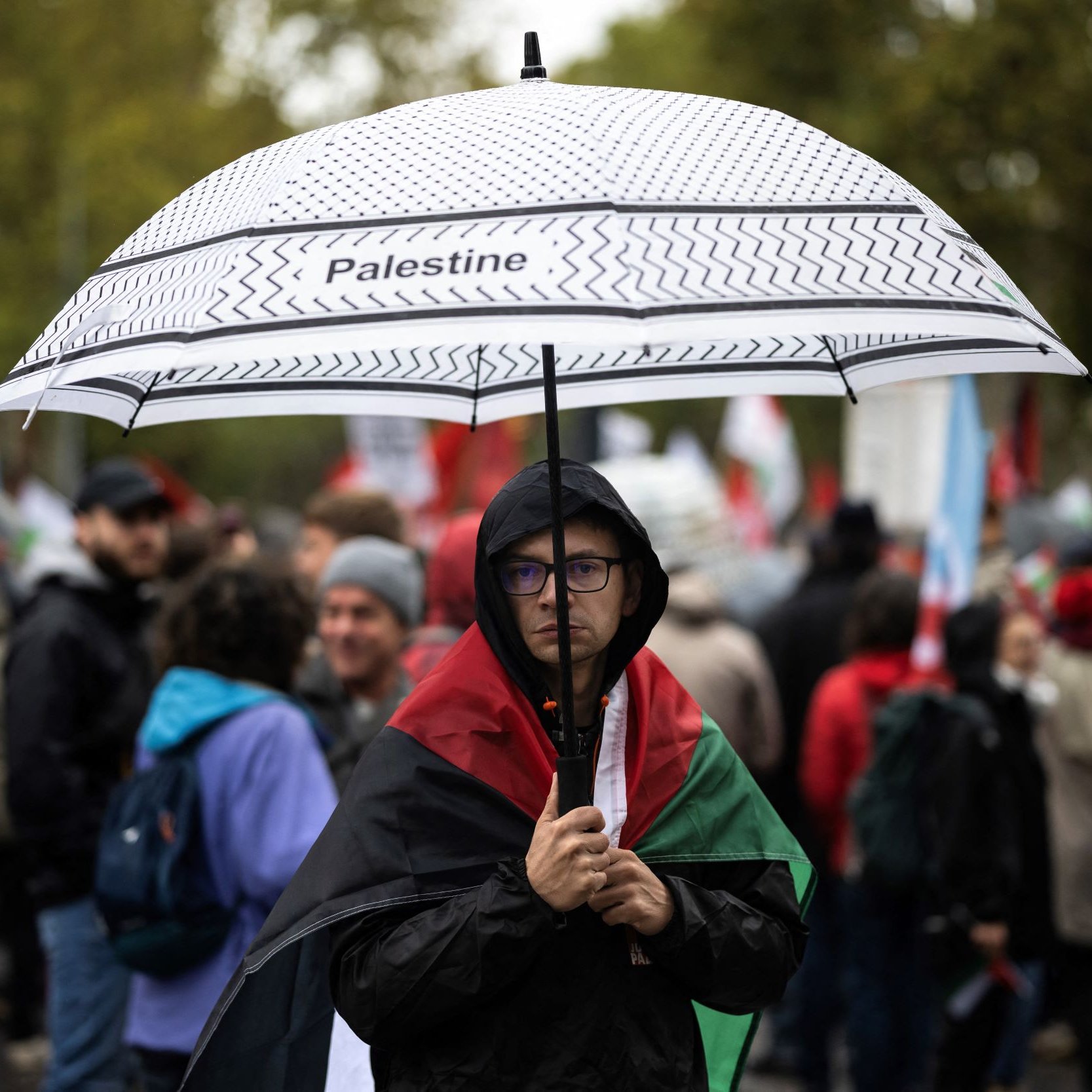 L'image montre une personne portant un parapluie noir et blanc sur lequel est écrit "Palestine". Cette personne est habillée d'un manteau sombre et se tient au milieu d'une foule qui semble participer à une manifestation. En arrière-plan, on peut apercevoir d'autres manifestants, certains portant des drapeaux et des pancartes. L'ambiance semble sérieuse et déterminée, avec des arbres et un ciel nuageux en arrière-plan.