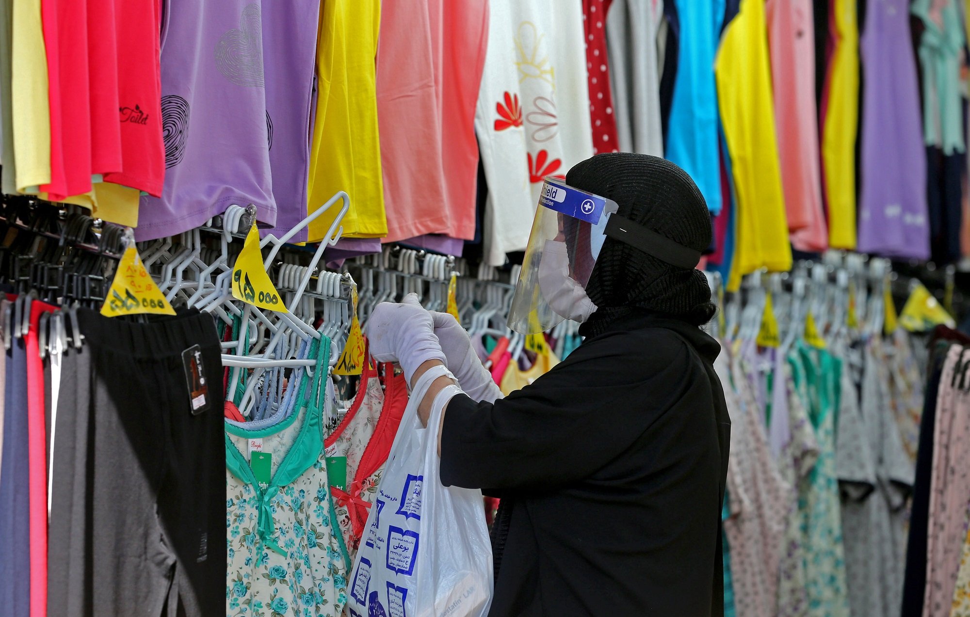 The image depicts a person wearing a black hijab and a face shield, shopping for clothing in a store. They are holding a white shopping bag and carefully examining different tops displayed on hangers. The store features a colorful array of clothing items, including various shades of purple, yellow, and floral patterns, hanging on racks in the background. The environment suggests a retail space focused on apparel, likely catering to a diverse range of styles.