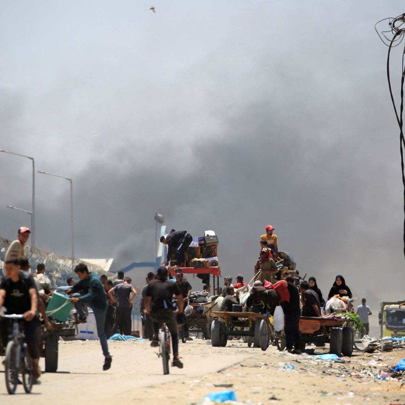 L'immagine mostra una scena di un'area urbana colpita da un conflitto o una crisi. Si vedono diverse persone che si muovono lungo una strada, alcune in bicicletta, altre a bordo di carri trainati da animali. Sullo sfondo, si nota del fumo denso che si alza, suggerendo un incendio o una situazione pericolosa. Le persone sembrano essere coinvolte in attività di trasporto, mentre l'atmosfera è tesa e caotica, con una presenza significativa di detriti e rifiuti sulla strada.