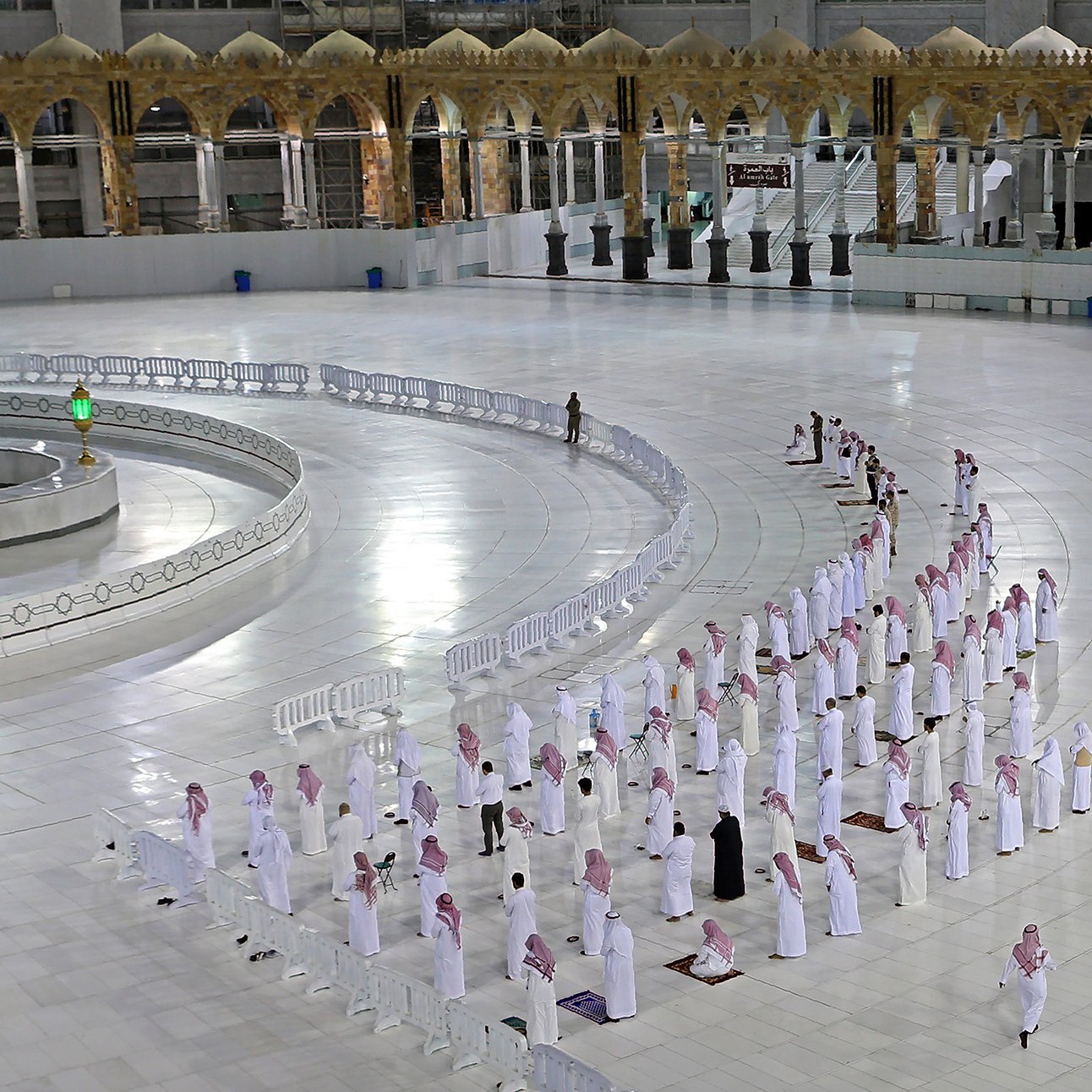 La imagen muestra el interior de la Gran Mezquita de La Meca, donde se encuentra la Kaaba. Se puede ver un gran espacio blanco con filas de personas en posición de oración. Están vistiendo tradicionalmente con túnicas blancas y pañuelos en la cabeza. En el fondo, se aprecia la Kaaba con su cubierta negra y algunos elementos decorativos. También hay barreras que delimitan el espacio y un par de agentes de seguridad en el área. La atmósfera es de calma y devoción.
