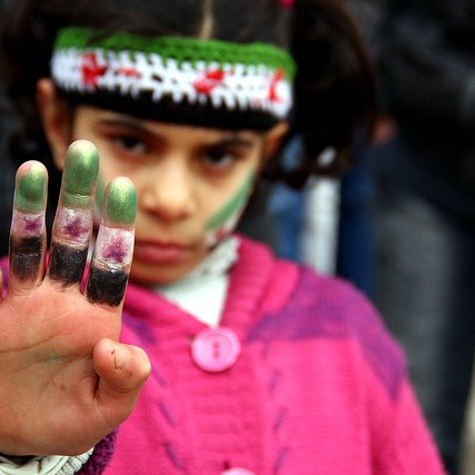 L'image montre une jeune fille qui lève la main en avant, exhibant ses doigts teintés de peinture. Elle porte un pull en laine rose et un bandeau coloré orné de motifs. Son visage est marqué par des traces de peinture, notamment sur les joues. L'expression de son visage semble déterminée, et elle attire l'attention sur son geste. En arrière-plan, on peut apercevoir des silhouettes floues de personnes, suggérant un rassemblement ou une manifestation.