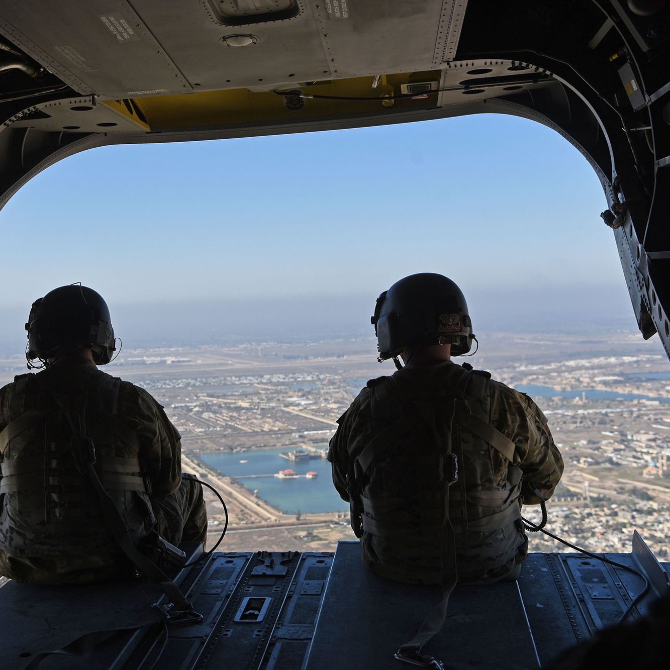 در این تصویر، دو نفر با تجهیزات نظامی در حال نشستن در یک هلیکوپتر هستند. آن‌ها در حال مشاهده مناظر بیرون از هلیکوپتر هستند که نمایی از یک شهر و آب‌های اطراف آن قابل مشاهده است. آسمان آبی و روشنی روز نشان‌دهنده یک روز صاف است. محیط داخلی هلیکوپتر نیز شامل تجهیزات و سیم‌کشی‌های نظامی است.