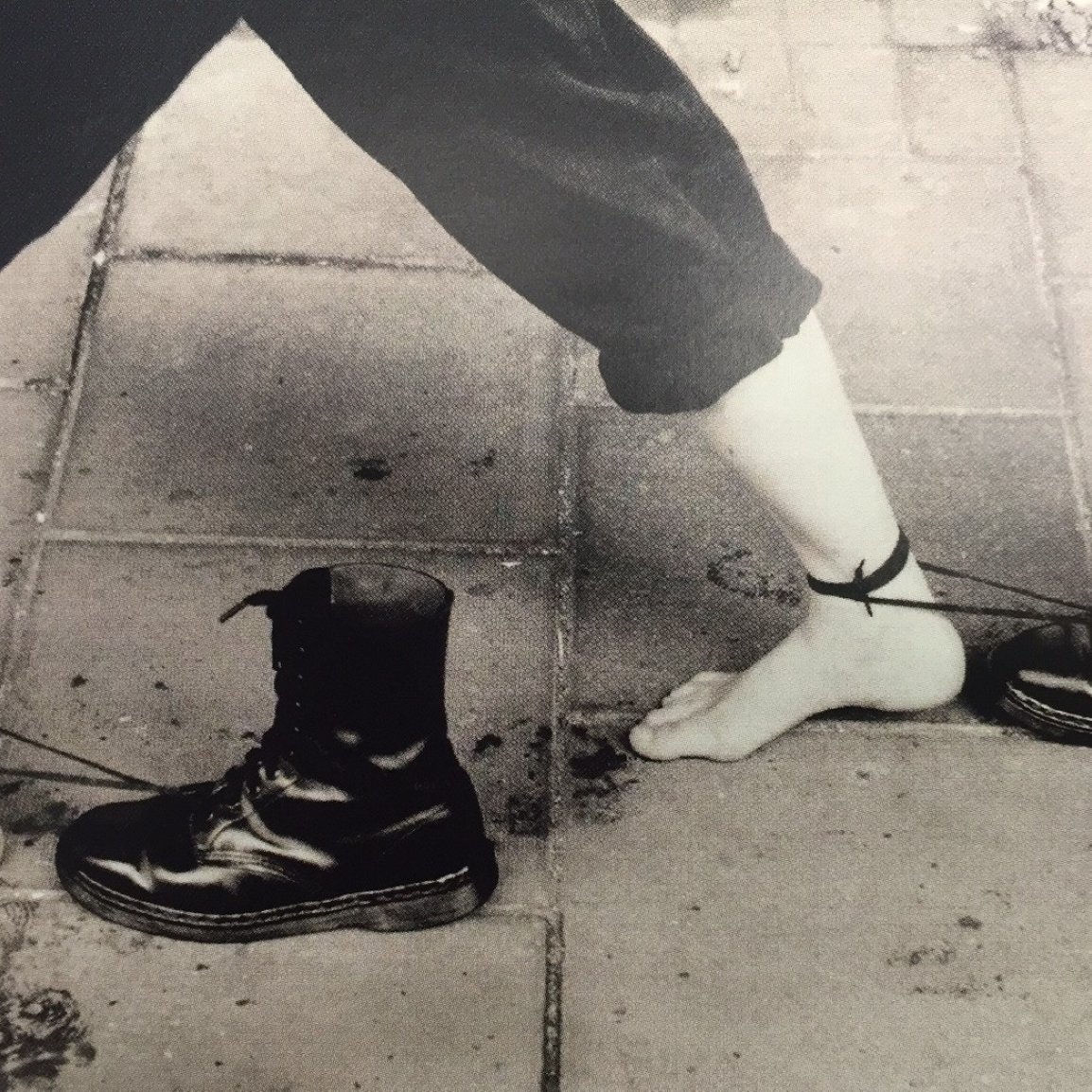 Des pieds nus liés à des bottes noires par des cordes. Image en noir et blanc.