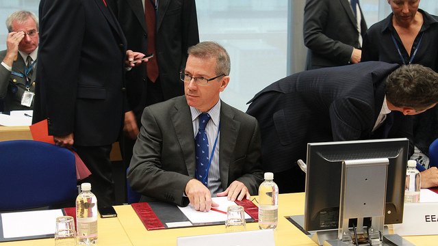 L'image montre une scène de réunion où plusieurs personnes sont présentes autour d'une table. Un homme, assis au premier plan, porte un costume gris et une cravate bleue à pois. Il semble concentré, en train d'organiser des documents devant lui. Sur la table, il y a des bouteilles d'eau et des documents disposés. À l'arrière-plan, d'autres participants sont visibles, certains discutant ou se déplaçant. L'environnement semble formel, typique d'une conférence ou d'une réunion professionnelle.