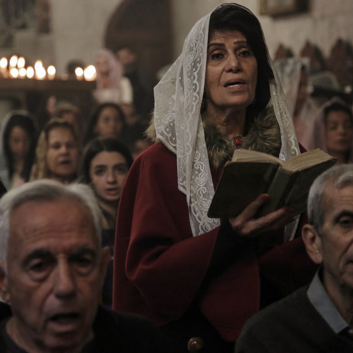 L'immagine mostra un gruppo di persone durante una cerimonia, probabilmente in una chiesa. Una donna al centro, che indossa un velo bianco e un abito rosso, tiene in mano un libro e sembra partecipare attivamente alla celebrazione. Intorno a lei ci sono altre persone, alcune delle quali appaiono in preghiera o cantando. L'atmosfera è solenne, con un gioco di luci morbide e candele accese sullo sfondo che creano un'illuminazione calda e accogliente.