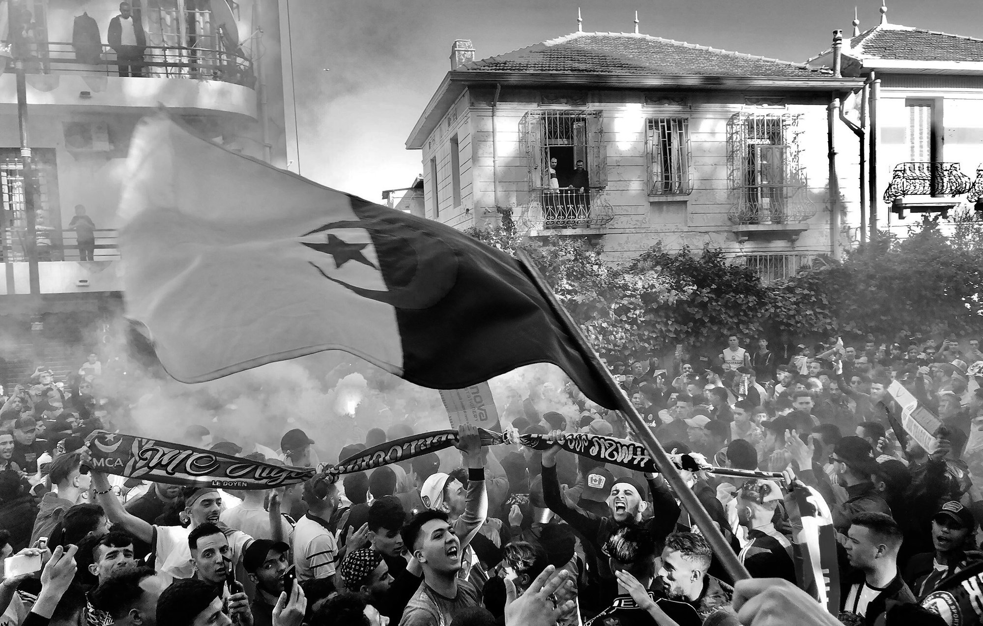 The image depicts a large crowd gathered in an urban setting, likely during a festive or protest event. People are seen waving flags and holding scarves, creating a vibrant atmosphere. Smoke is rising in the air, possibly from flares or fireworks, adding to the intensity of the scene. The buildings in the background have a mix of architectural styles, hinting at a lively neighborhood. The overall mood appears energetic and spirited, with individuals showing strong emotions and camaraderie.