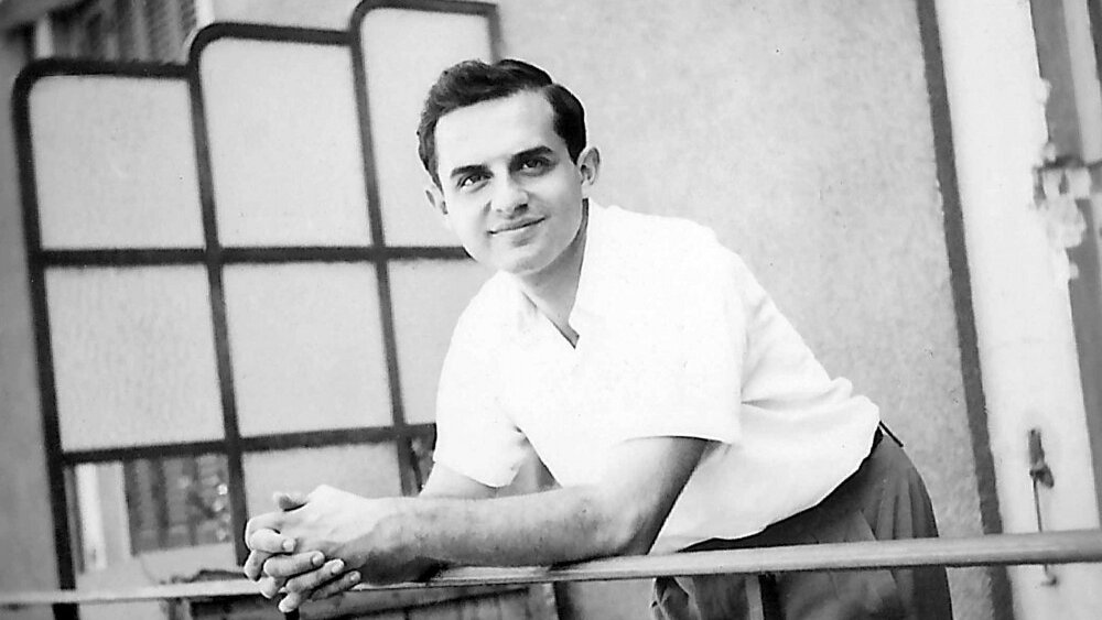 The image features a young man with a pleasant expression, leaning on a railing. He has dark hair styled in a neat manner and is dressed in a light-colored short-sleeve shirt paired with trousers. The background suggests a balcony or outdoor setting, with a partially visible modern-style partition or screen. The photo has a vintage quality, indicating it was likely taken several decades ago.