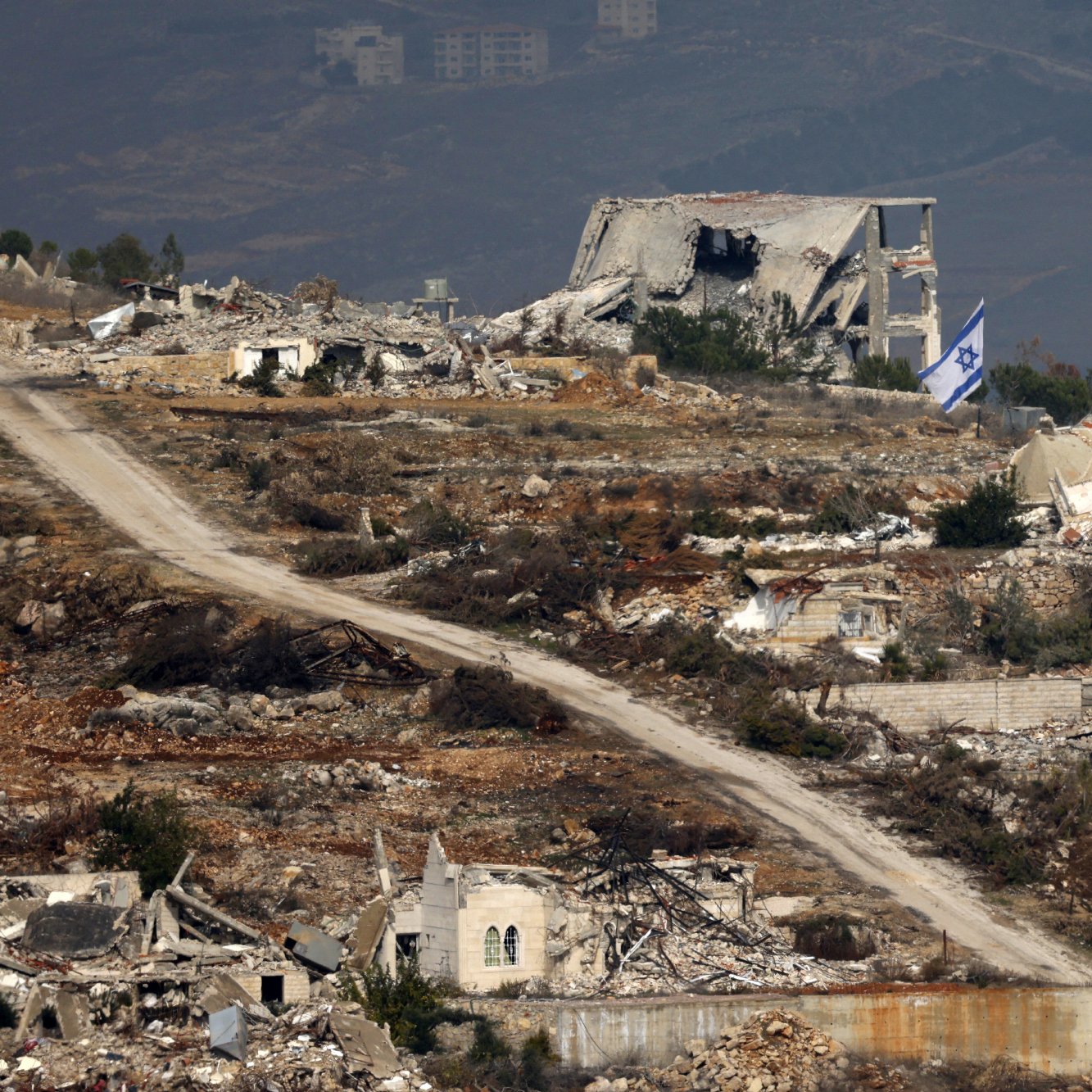 The image depicts a desolate landscape featuring extensive destruction, with several ruined buildings scattered throughout the area. In the foreground, there is a dirt road winding through the wreckage. Among the ruins, an Israeli flag can be seen prominently displayed, suggesting a connection to the location. The background shows additional structures and what appears to be a mountainous or hilly terrain, indicating a possibly rural setting. The overall scene conveys a sense of abandonment and turmoil.