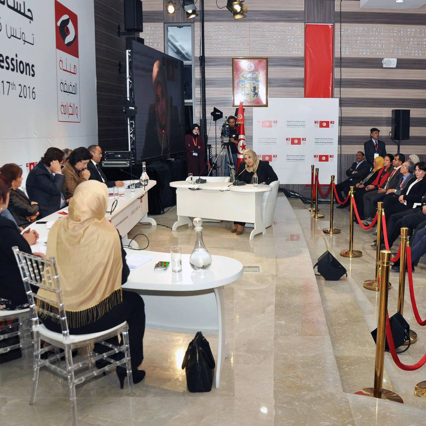 الصورة تظهر جلسة استماع تتعلق بموضوع اجتماعي أو سياسي. في المكان، يجلس عدد من الأشخاص على الجانبين، حيث يبدو أن هناك مجموعة من المتحدثين يجلسون على منصة مرتفعة. يظهر أمامهم طاولة مستديرة حيث يتم تقديم المداخلات. تُظهر الخلفية شعارات وصورة لمتحدث ما، مما يشير إلى أهمية الحدث. الأجواء تبدو رسمية ومرتبة، مع وجود حواجز حمراء تفصل بين الحضور.
