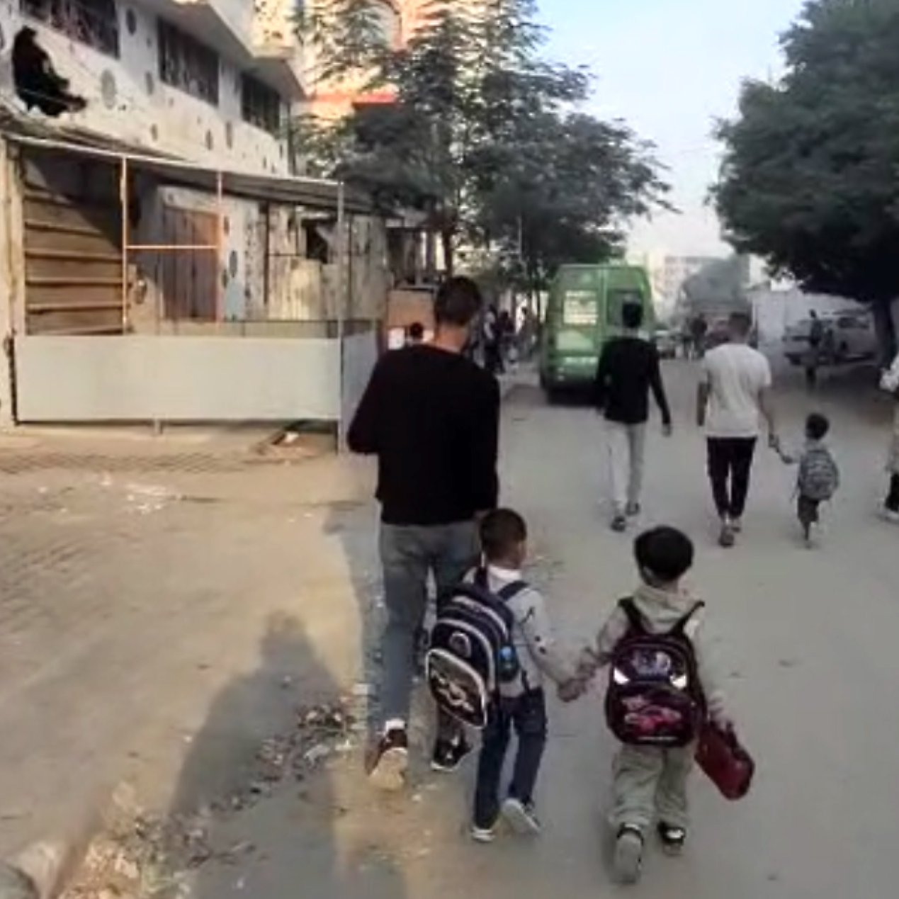 Un uomo cammina con due bambini, mentre altre persone si muovono in una strada cittadina.
