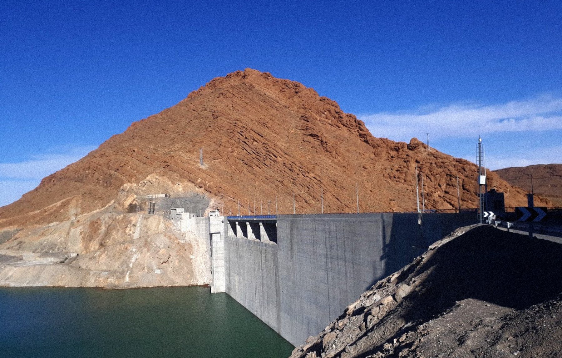 La imagen muestra un gran embalse o represa construida de concreto, situada a la orilla de un cuerpo de agua de color verde. Detrás de la represa se puede ver una montaña de tonalidades marrones y naranjas, con un cielo despejado de un azul brillante en el fondo. La estructura de la represa es sólida y parece estar bien mantenida, mientras que el terreno alrededor tiene un aspecto árido y rocoso.