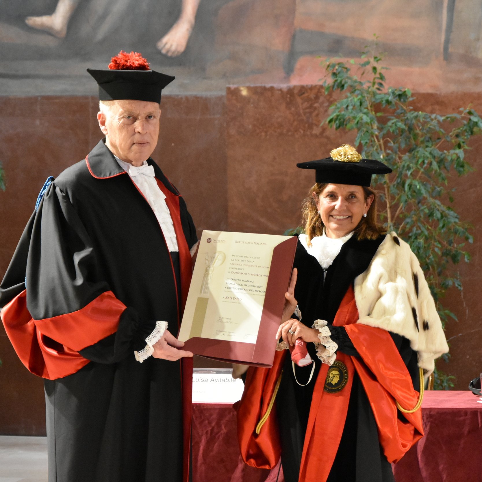 L'immagine mostra due persone vestite in abiti accademici formali, tipici per cerimonie di laurea o conferimenti di titoli. Il soggetto a sinistra indossa un mantello nero con dettagli rossi e un cappello, mentre a destra c'è una donna con un abito simile, ma con un mantello e un cappello che contengono diversi elementi decorativi. Entrambi stanno tenendo un documento o un certificato che sembra essere di rilevanza accademica o ufficiale. Sullo sfondo, ci sono piante e un muro decorato, indicando un ambiente formale e celebrativo.