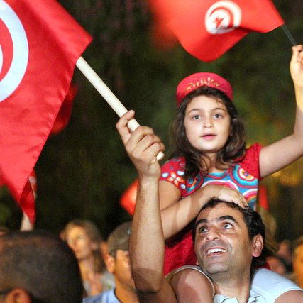 L'image montre une scène de célébration en Tunisie. Une jeune fille, vêtue d'une robe colorée, est assise sur les épaules d'un homme qui sourit. Ils brandissent des drapeaux tunisiens. En arrière-plan, on aperçoit une foule joyeuse, créant une atmosphère festive. Les expressions des visages reflètent la joie et l'engagement.