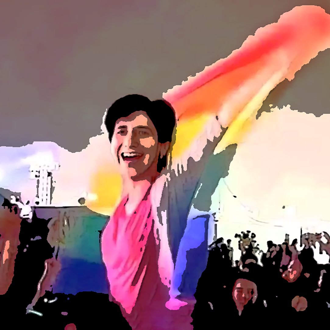 En la imagen se puede ver a una persona sonriente que sostiene una bandera de colores del arcoíris, símbolo del movimiento LGBTQ+. Está en un ambiente festivo, probablemente en un evento o celebración, rodeada de una multitud que también parece estar disfrutando del momento. La escena transmite una sensación de alegría y celebración de la diversidad.