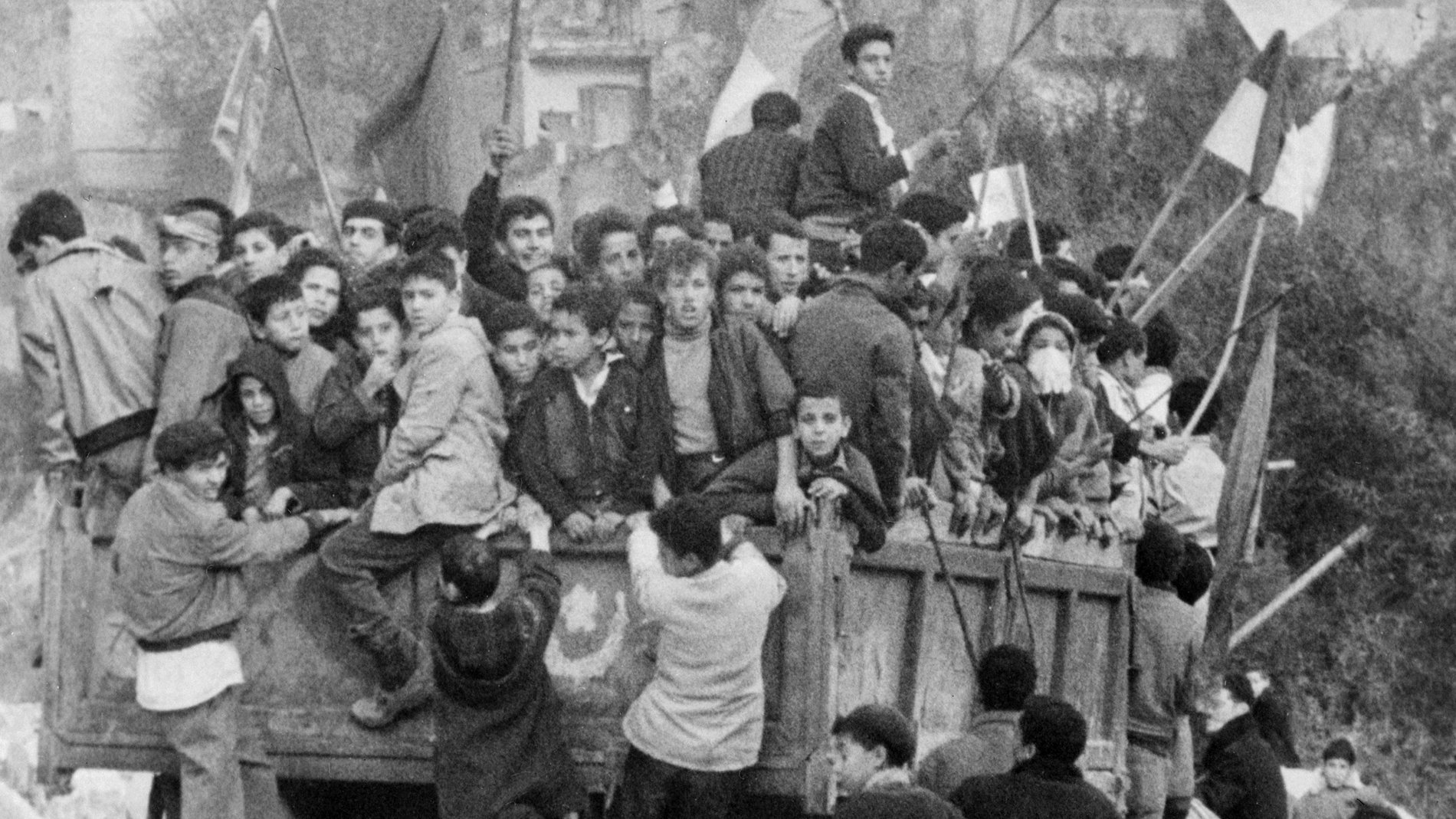 L'image montre un groupe de jeunes garçons et d'adolescents rassemblés sur un camion, entourés de drapeaux. Ils semblent célébrer un événement, peut-être une manifestation ou une fête. Les enfants sont penchés et accrochés au camion, reflétant un moment de camaraderie ou d'excitation collective. En arrière-plan, on aperçoit des bâtiments typiques d'une zone urbaine, contribuant à l'ambiance dynamique de la scène.
