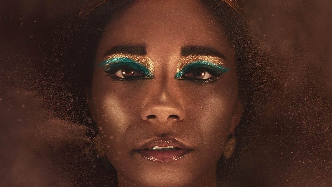 L'immagine presenta un volto femminile con un trucco molto elaborato, caratterizzato da ombretti blu e oro che mettono in risalto gli occhi. La persona indossa una corona decorata con serpenti dorati, che conferisce un aspetto regale e misterioso. Lo sfondo è sfocato e sembra essere avvolto da una polvere dorata, creando un'atmosfera affascinante e drammatica. La luce è calda e accentua la bellezza del viso, rendendo l'immagine suggestiva e intrigante.