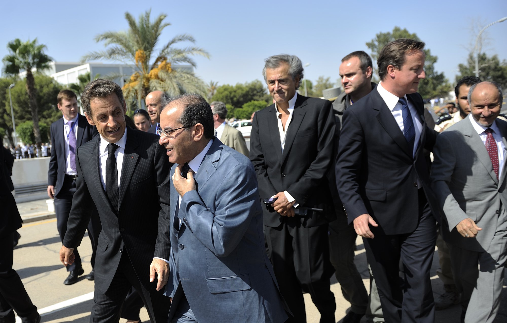 L'image montre un groupe d'hommes marchant ensemble dans un environnement extérieur. Ils semblent être en discussion animée. En arrière-plan, on peut apercevoir des palmiers et un bâtiment, suggérant un climat ensoleillé. Les hommes portent des costumes, ce qui indique un contexte formel ou professionnel. L'ambiance semble dynamique et engageante.