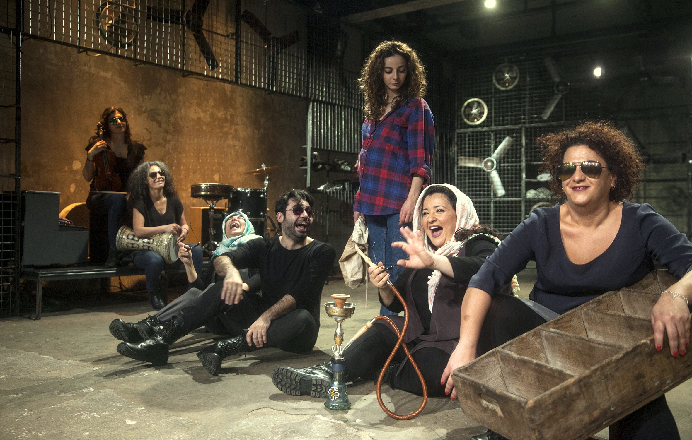 L'image présente un groupe de personnes assises sur le sol d'un espace décoratif et artistique. On y voit des femmes et des hommes, certains souriant et riant, dans une ambiance décontractée et joyeuse. Les femmes portent des vêtements variés, y compris un hijab, tandis que certains d'entre eux portent des lunettes de soleil. À l'arrière-plan, il y a des instruments de musique et des éléments de décor industriels, ce qui ajoute une touche créative à la scène. Au centre, une femme tient une chicha (narguilé) et semble engagée dans la conversation. L'ensemble de la composition donne une impression de camaraderie et de convivialité.