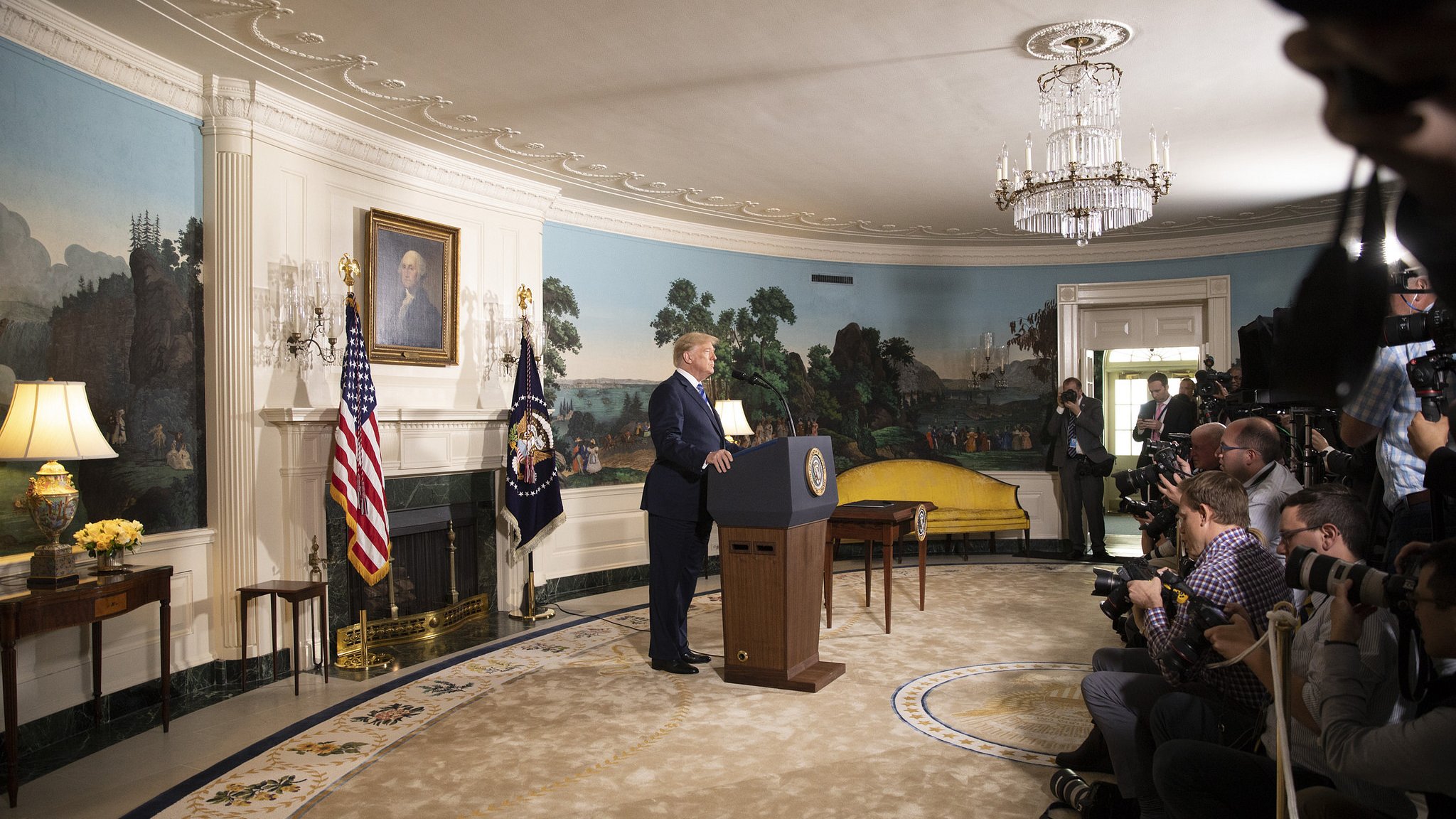 L'image montre un homme debout devant un podium, probablement lors d'une allocution publique. Il est situé dans une pièce décorée avec des murs peints de paysages, et des drapeaux américain et présidentiel sont visibles à côté de lui. Plusieurs journalistes et photographes sont présents, capturant l'événement. Le mobilier de la pièce semble ancien, et il y a un grand lustre au plafond.