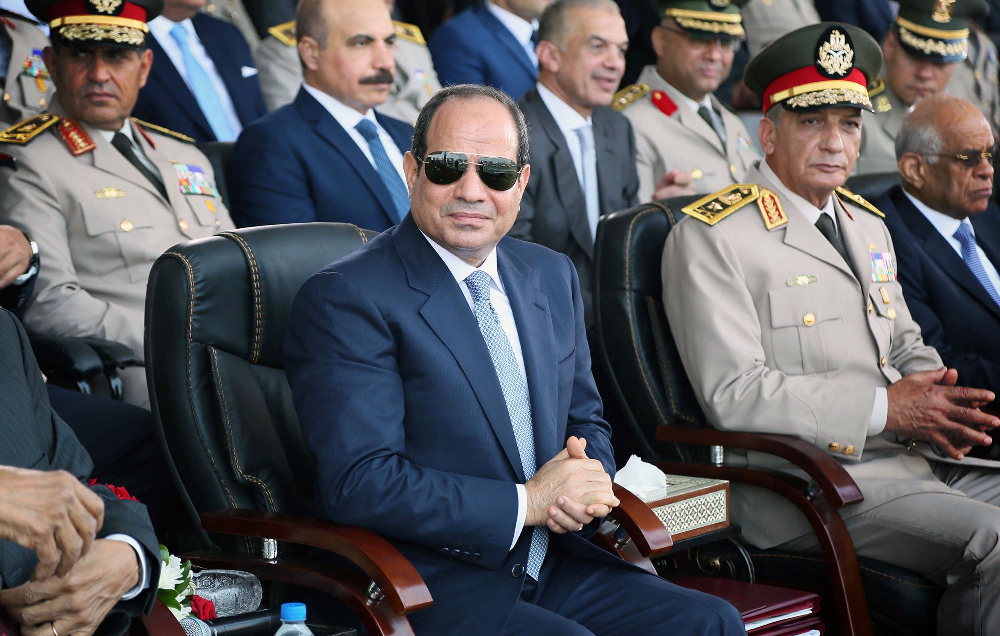 L'image montre un groupe de personnes lors d'un événement officiel. Au premier plan, un homme portant des lunettes de soleil et habillé d'un costume sombre est assis sur une chaise, semblant attentif. Il y a d'autres hommes en uniforme militaire et en tenue formelle derrière lui, qui semblent également être présents pour l'événement. L'ambiance est solennelle et il semble s'agir d'une cérémonie ou d'une célébration officielle.