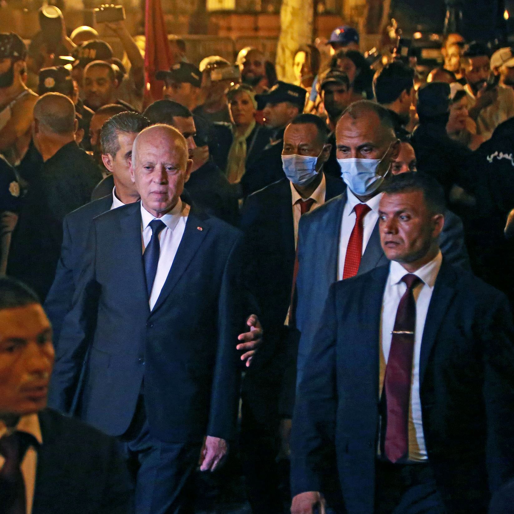 در این تصویر، گروهی از افراد در یک محیط شلوغ و پرجمعیت به‌نظر می‌رسند. در مرکز تصویر، فردی با کت و شلوار تیره در حال عبور است و چندین محافظ یا همراه در اطراف او حضور دارند. برخی از افراد در زمینه تصویر، ماسک به‌صورت دارند و دیگران مشغول تماشا و عکاسی از صحنه هستند. محیط به نظر می‌رسد شبانه باشد و نورها به‌خوبی در تصویر مشاهده می‌شوند.